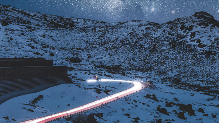 Night time landscape showing a snowing mountain and a road with car lights on it